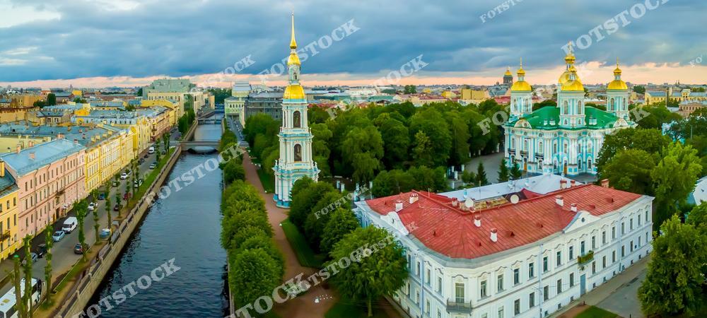 На фотографии изображен Никольский морской собор с его золотыми куполами, расположенный в окружении зелени. Рядом находится изящная Колокольня, стоящая на берегу канала. Утренний свет и облачное небо придают фотографии мягкую и спокойную атмосферу.